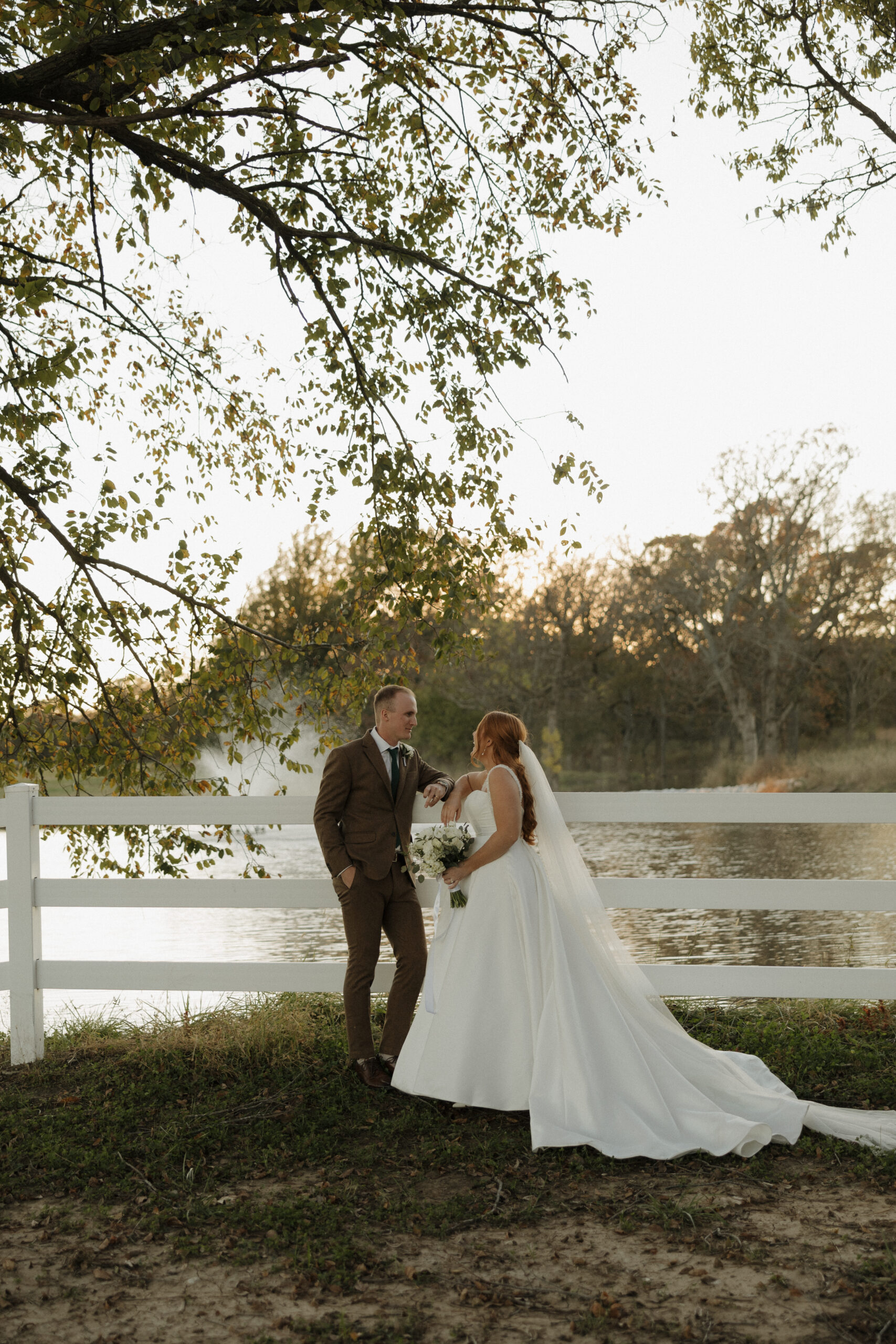 Bride and groom at Bluestem Manor wedding venue in Sperry, OK, posing by the water and white fence during a golden hour sunset session.
