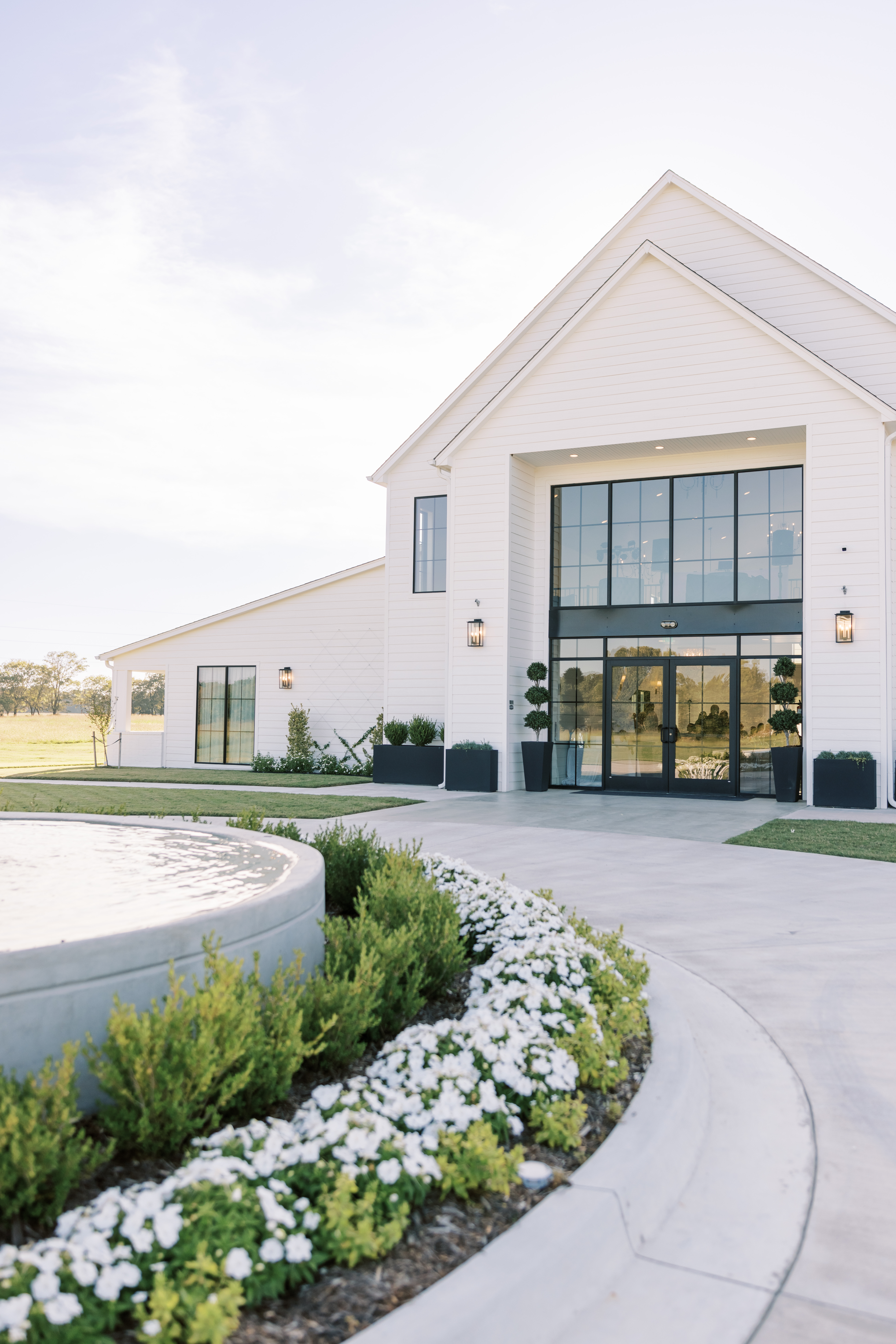 The grand entrance of Bluestem Manor, a modern white barn venue in Oklahoma, featuring large black-framed windows, a circular concrete driveway with a fountain, and white floral landscaping.