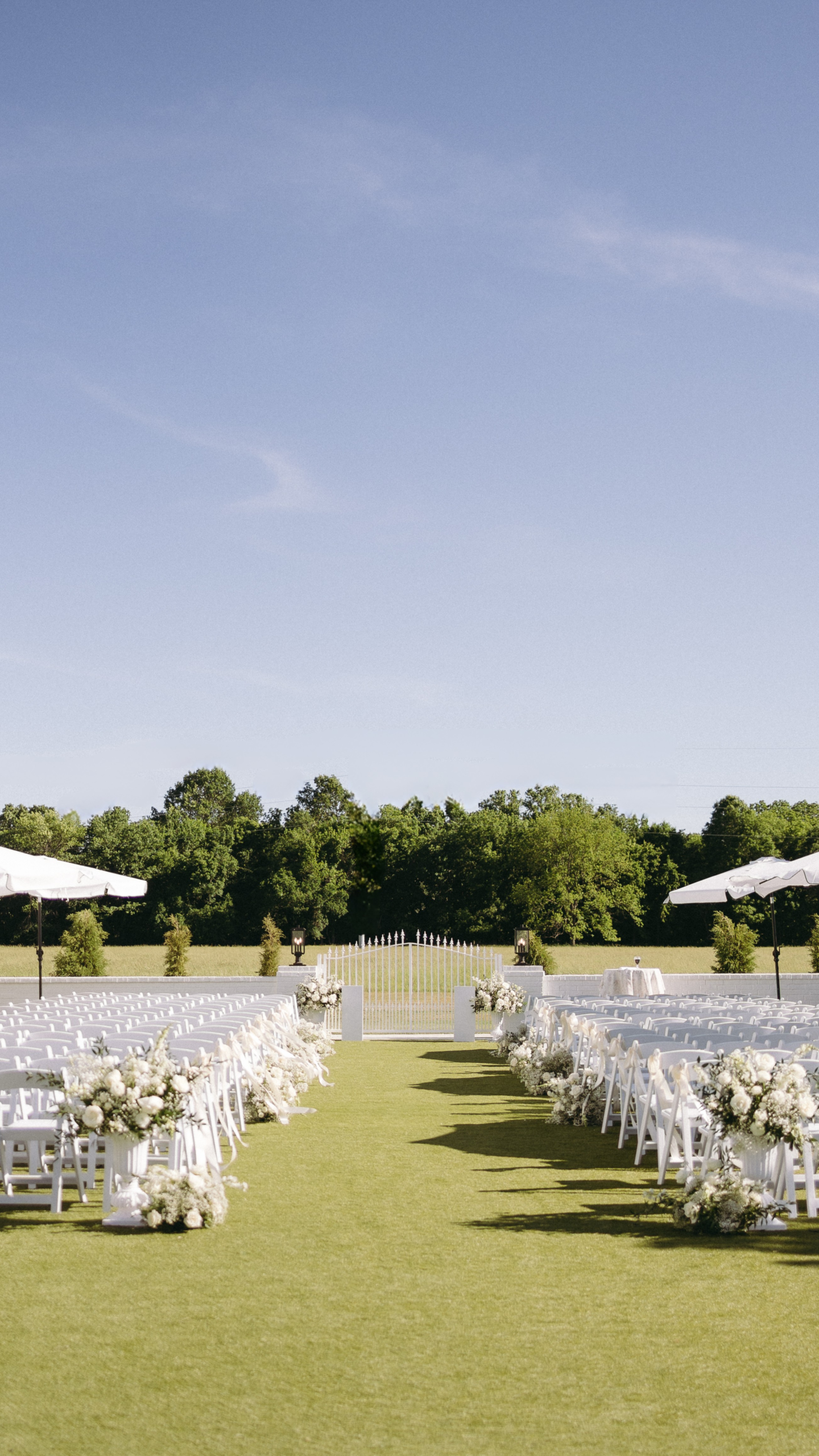 An outdoor wedding ceremony setup at Bluestem Manor featuring white chairs, floral aisle arrangements, and a white garden gate overlooking a lush green field under a clear blue sky.