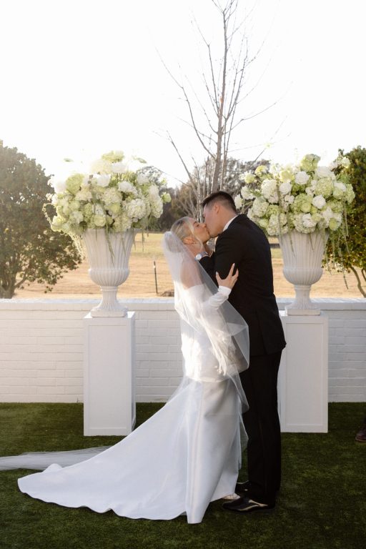 Newlyweds kissing between white floral urns during an outdoor wedding ceremony at Bluestem Manor.