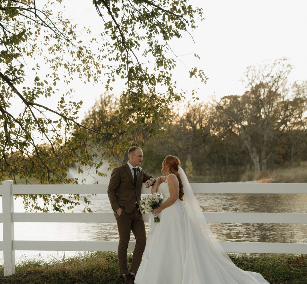 Bride and groom at Bluestem Manor wedding venue in Sperry, OK, posing by the water and white fence during a golden hour sunset session.